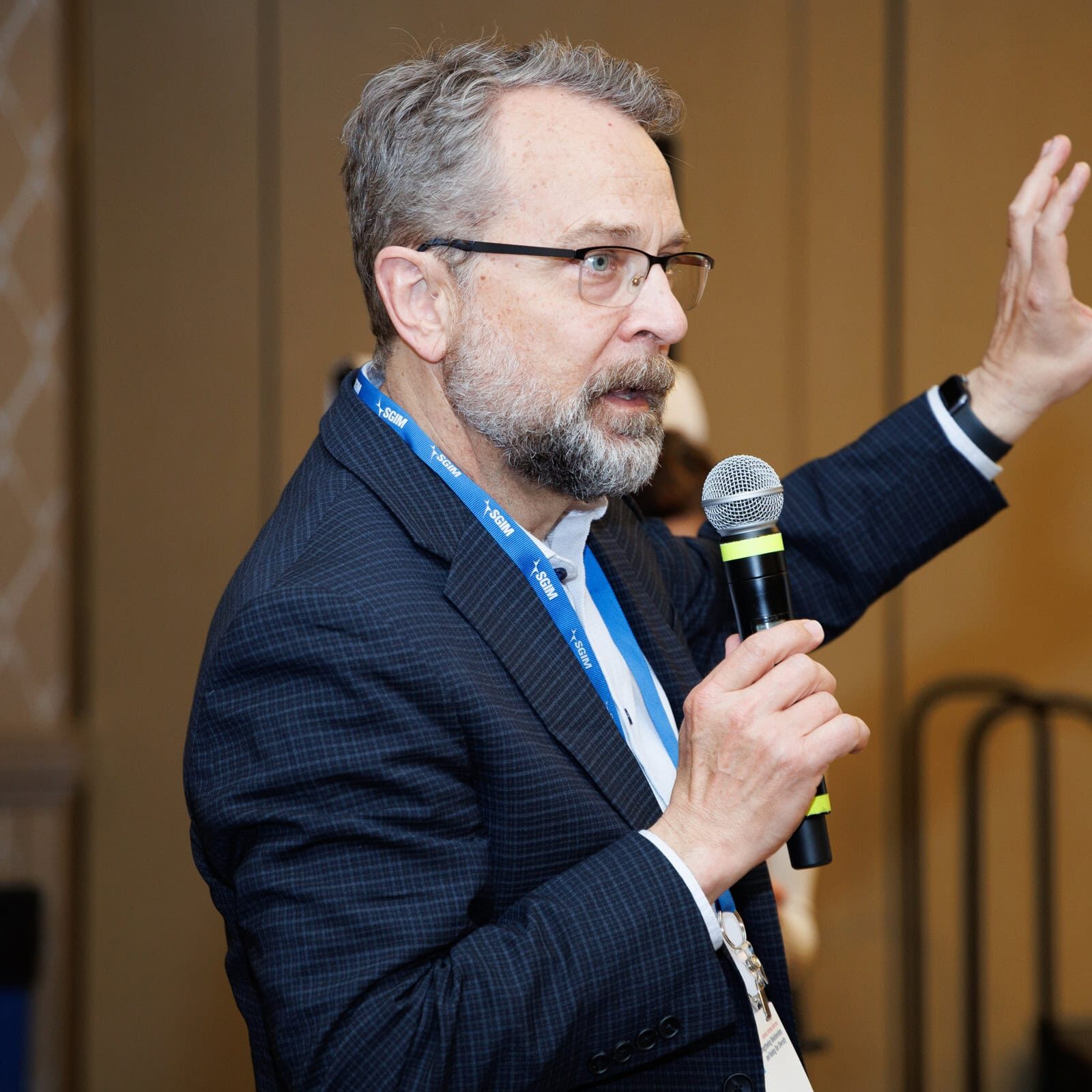 A man with glasses and a beard holding a microphone, speaking and gesturing with one hand during a presentation at SGIM23.