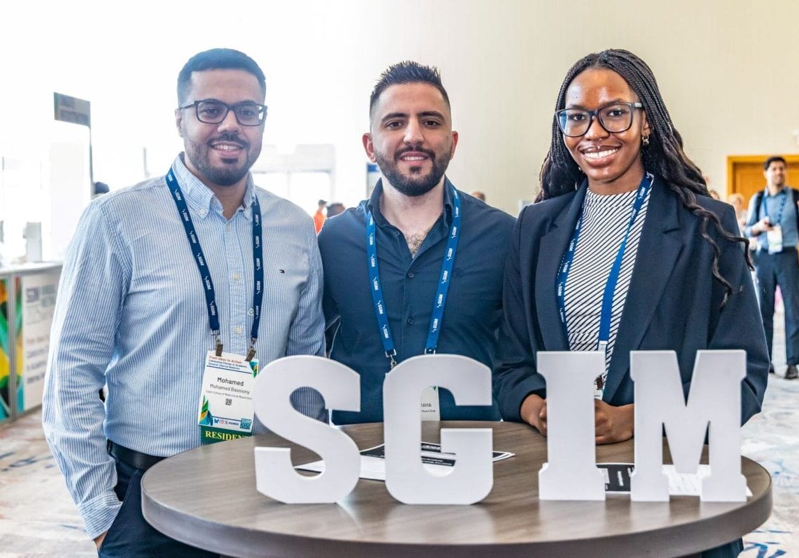 Three smiling conference attendees standing in front of letter blocks that read S.G.I.M.
