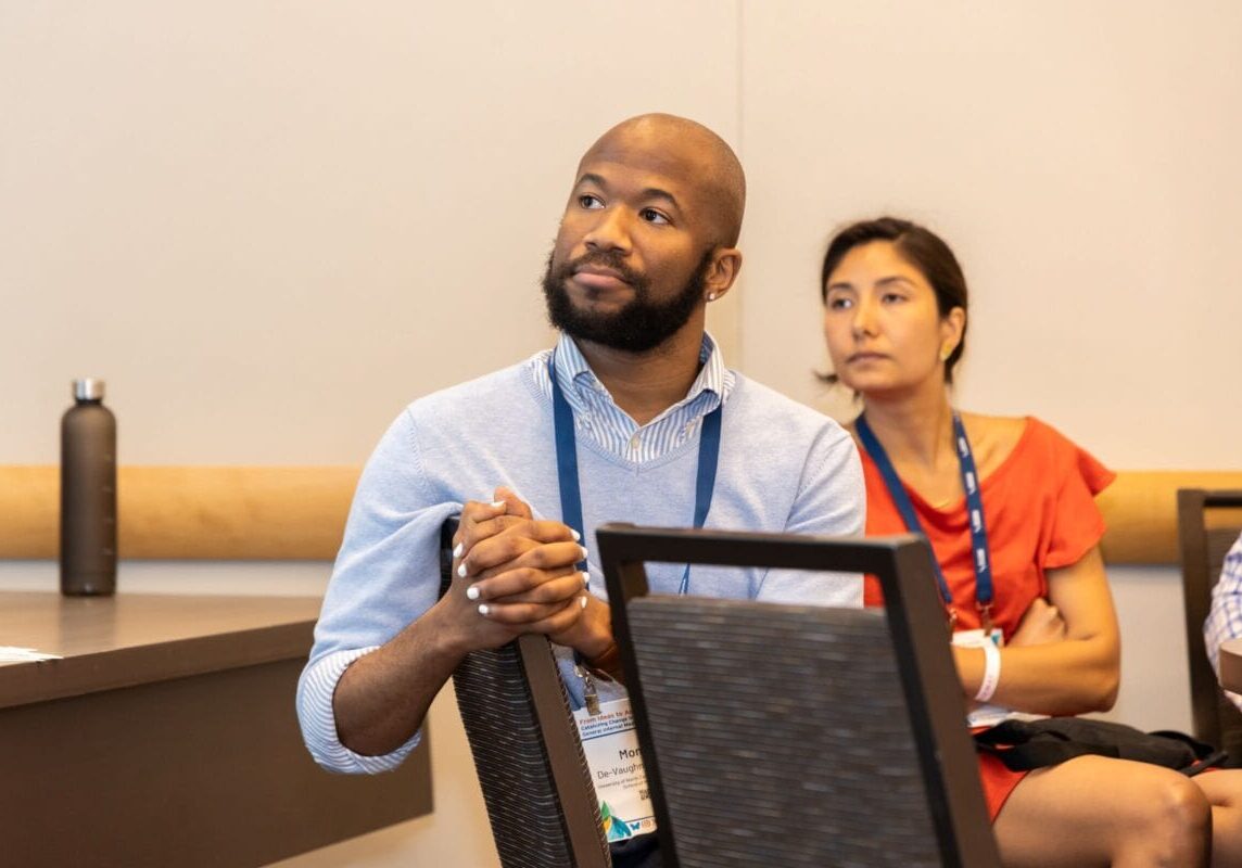 A man and woman sit in a conference room listening attentively, both wearing blue lanyards