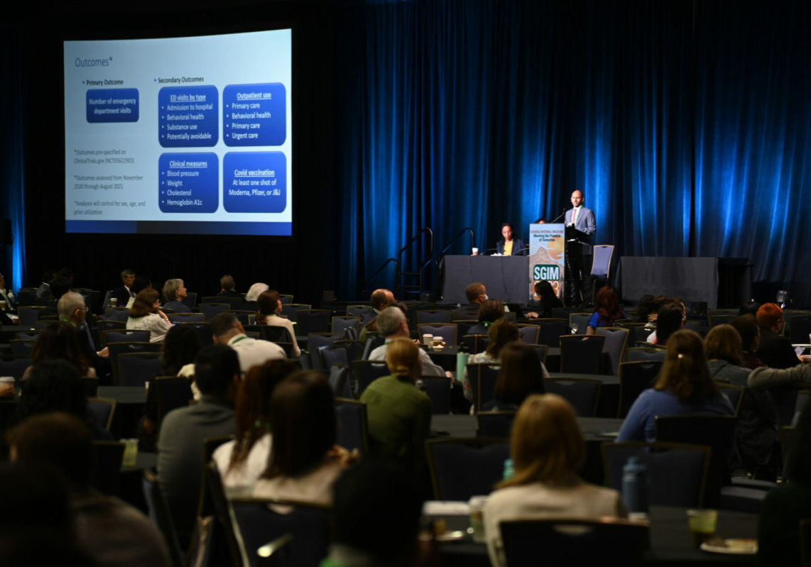 The image shows a large conference room filled with attendees seated and facing a stage where a speaker is presenting. The stage has two people seated at a table, and one person standing at a podium, addressing the audience. A large screen to the left of the stage displays a slide with text and diagrams related to the presentation.