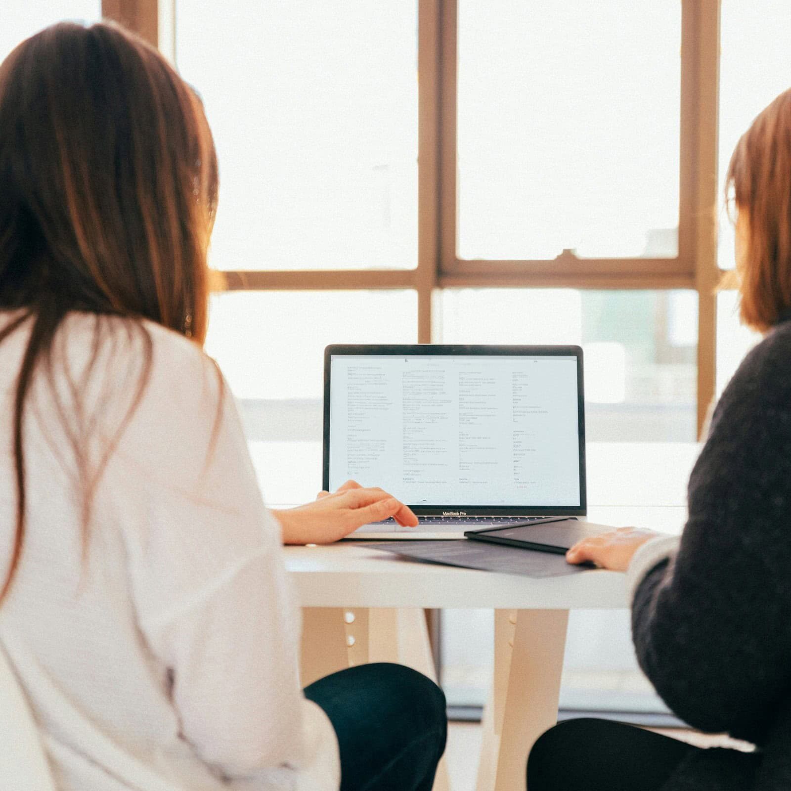 Two women sitting at a table, working together on a laptop with a website open on the screen in a bright, modern office setting.