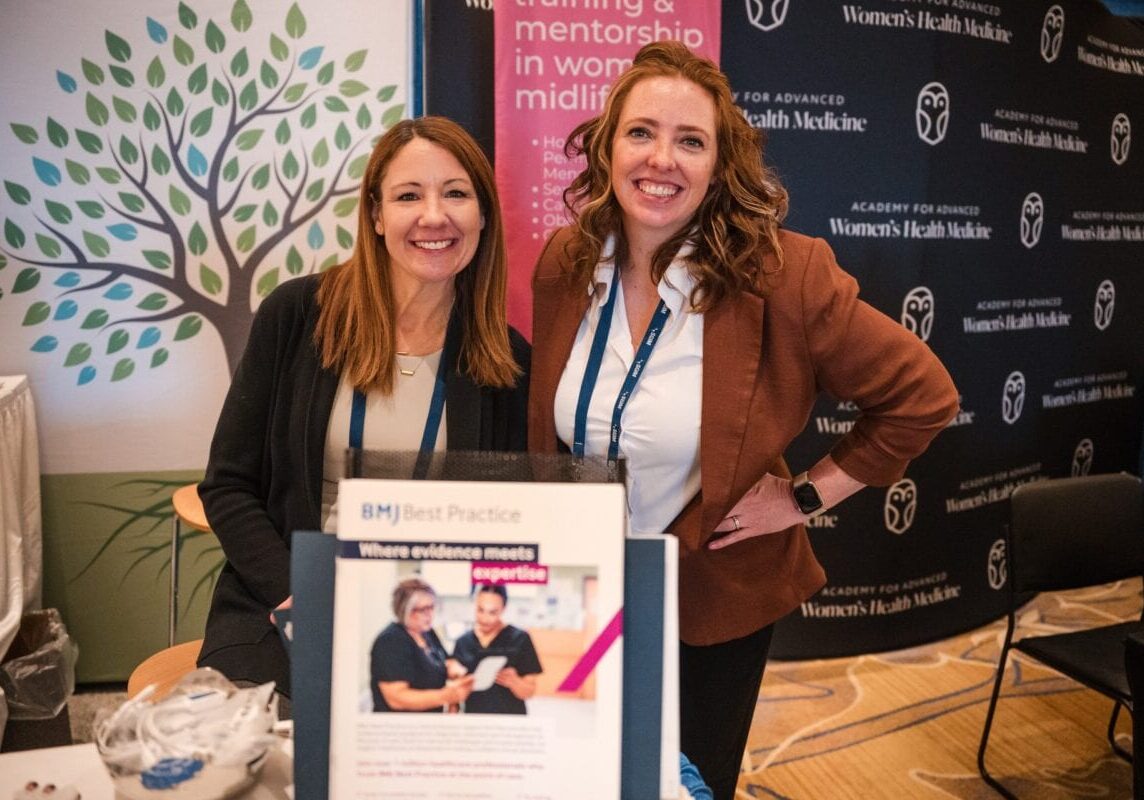 Two women smiling at an exhibitor booth with health-education banners behind them.