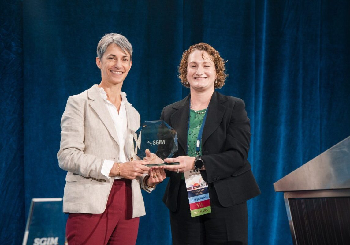 Two women on stage holding an SGIM-branded glass award, smiling toward the camera.